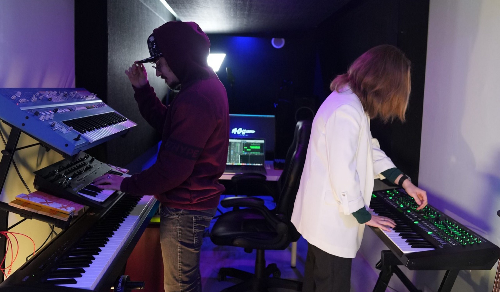 Two musicians at work in the room: one at a stack of synthesisers along the left wall, the other at a keyboard on the right, the desk and its screen between them.