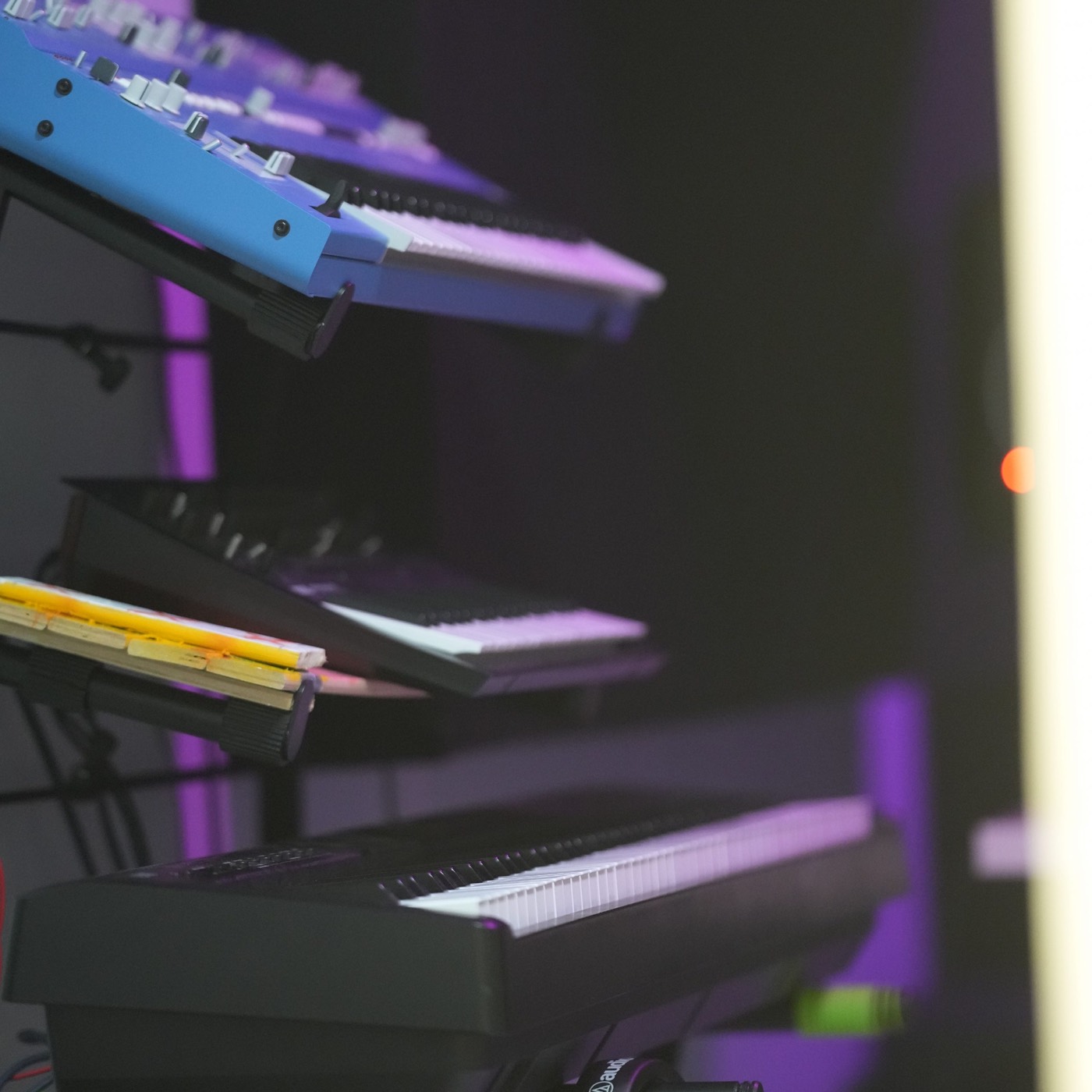 A tiered stand of synthesisers and keyboards in the studio, lit violet.