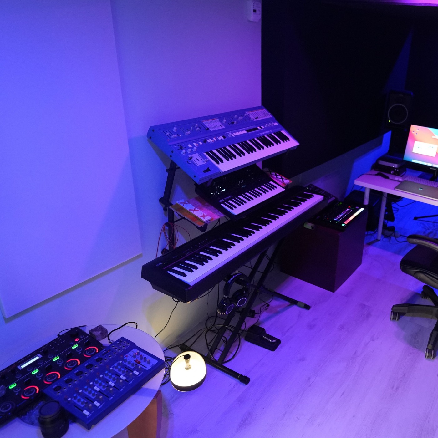 The studio's keyboard corner in blue and violet light: synthesisers stacked on a stand, a combo amp, and the desk with its screen behind.